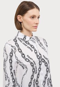 Άσπρη πουκάμισα με κουμπιά, με σχέδιο αλυσίδας και κείμενο "Versace Jeans Couture" σε μαύρο χρώμα. Φτιαγμένο από απαλό, ελαφρύ ύφασμα.