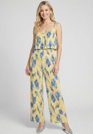 Femme souriante aux longs cheveux blonds portant une combinaison sans manches jaune à motifs floraux et des talons argentés, debout devant un arrière-plan gris uni.