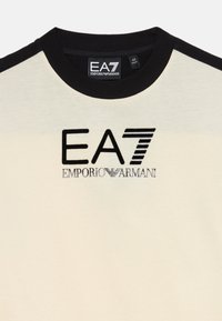 Trumpas, kremiškos spalvos marškinėliai su juodu apykaklėje ir rankovėmis. Priekyje yra išsiuvinėti "EA7" ir "EMPORIO ARMANI" logotipai.