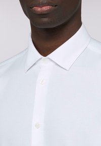 Camicia bianca con colletto in tessuto liscio, caratterizzata da una chiusura nascosta con bottoni trasparenti e un discreto dettaglio del logo sul lato sinistro.
