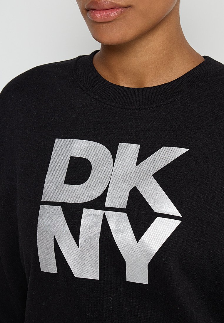 Nahaufnahme einer Person, die einen schwarzen Pullover mit großen silbernen Buchstaben "DKNY" auf der Vorderseite trägt.