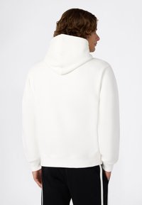 Uomo con capelli ricci che indossa un semplice hoodie bianco e pantaloni neri con strisce laterali bianche, in piedi con le spalle rivolte verso di noi, con le mani in tasca.