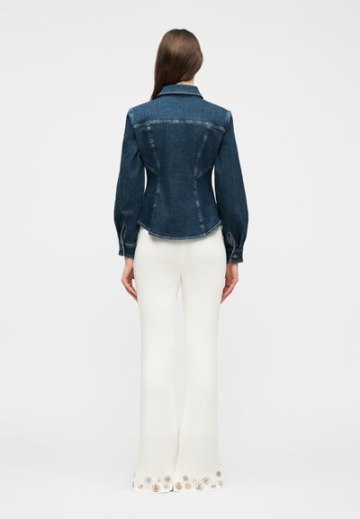 Γυναίκα που φαίνεται από πίσω, φοράει εφαρμοστή σκούρα τζιν ζακέτα και λευκό καμπανέ παντελόνι με ανθισμένες διακοσμήσεις στο τελείωμα.