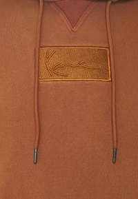 Hnedý mikina s jemnou textúrou, zdobená výšivkou s logom v písmene a kontrastnou šnúrkou s kovovými koncami.