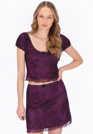 Tânără cu păr lung și ondulat, purtând un top crop cu mâneci scurte și imprimeu de leopard mov și o fustă asortată, stând cu mâna pe șold.