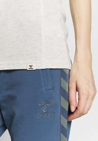 Lichtgrijze top gecombineerd met blauwe broek featuring een geborduurd logo en gestreepte zijkanten in groen en blauw. Glad stoftexturen.