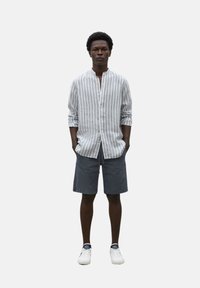Camisa de rayas de manga larga en blanco y azul, combinada con pantalones cortos grises. El modelo lleva zapatillas blancas con detalles oscuros y las manos en los bolsillos.