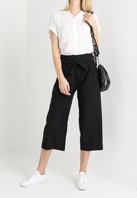 Witte blouse met korte mouwen en een notch kraag, gecombineerd met zwarte cropped broek met een gestrikt tailleband. Model draagt witte sneakers.