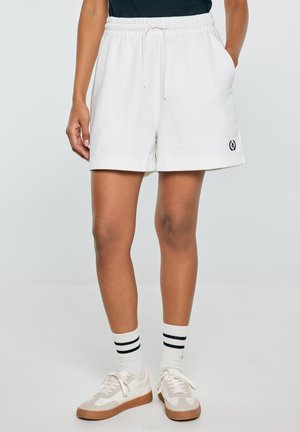 Osoba w białych szortach na sznurku, białych skarpetkach z dwoma czarnymi paskami oraz białych trampkach z beżowymi akcentami. Ręce częściowo w kieszeniach.