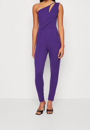 Kobieta ubrana w fioletowy kombinezon na jedno ramię z dopasowanymi nogawkami i czarnymi sandałami na wysokim obcasie z odkrytymi palcami, stojąca na jednolitym białym tle.
