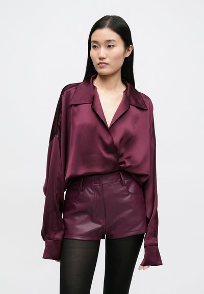 Bordo satino marškiniai su sagomis, apykakle ir ilgomis rankovėmis, derinami su aukštos juosmens bordo odinėmis šortais ir juodomis pėdkelnėmis.