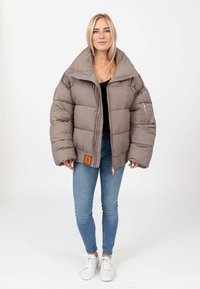 Giacca imbottita color marrone chiaro con un ampio colletto, chiusura con zip, e una tasca sulla manica. Presenta un'etichetta arancione in basso.