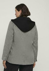 Donna con capelli corti e scuri che indossa una giacca grigia testurizzata con un grande cappuccio nero, vista di schiena su uno sfondo chiaro e uniforme.