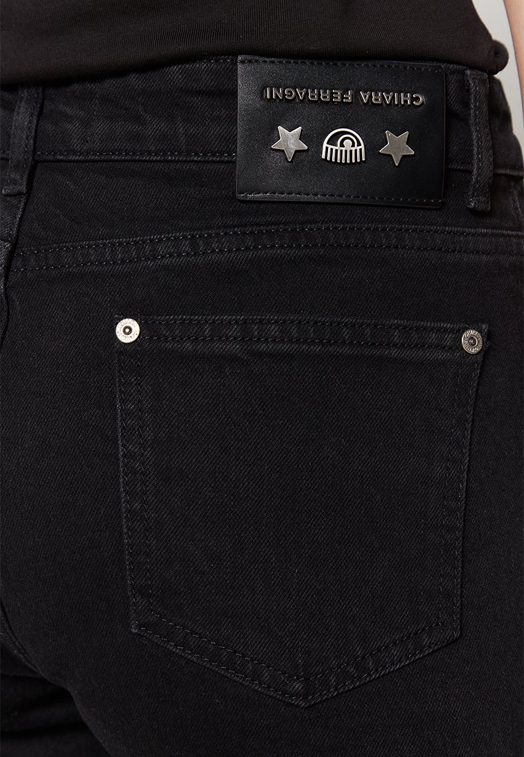 Schwarze Jeans aus Denim mit einem Lederetikett, das silberne Sterne akzentuiert, und einem stilisierten Regenbogen-Design, wobei eine Gesäßtasche sichtbar ist.