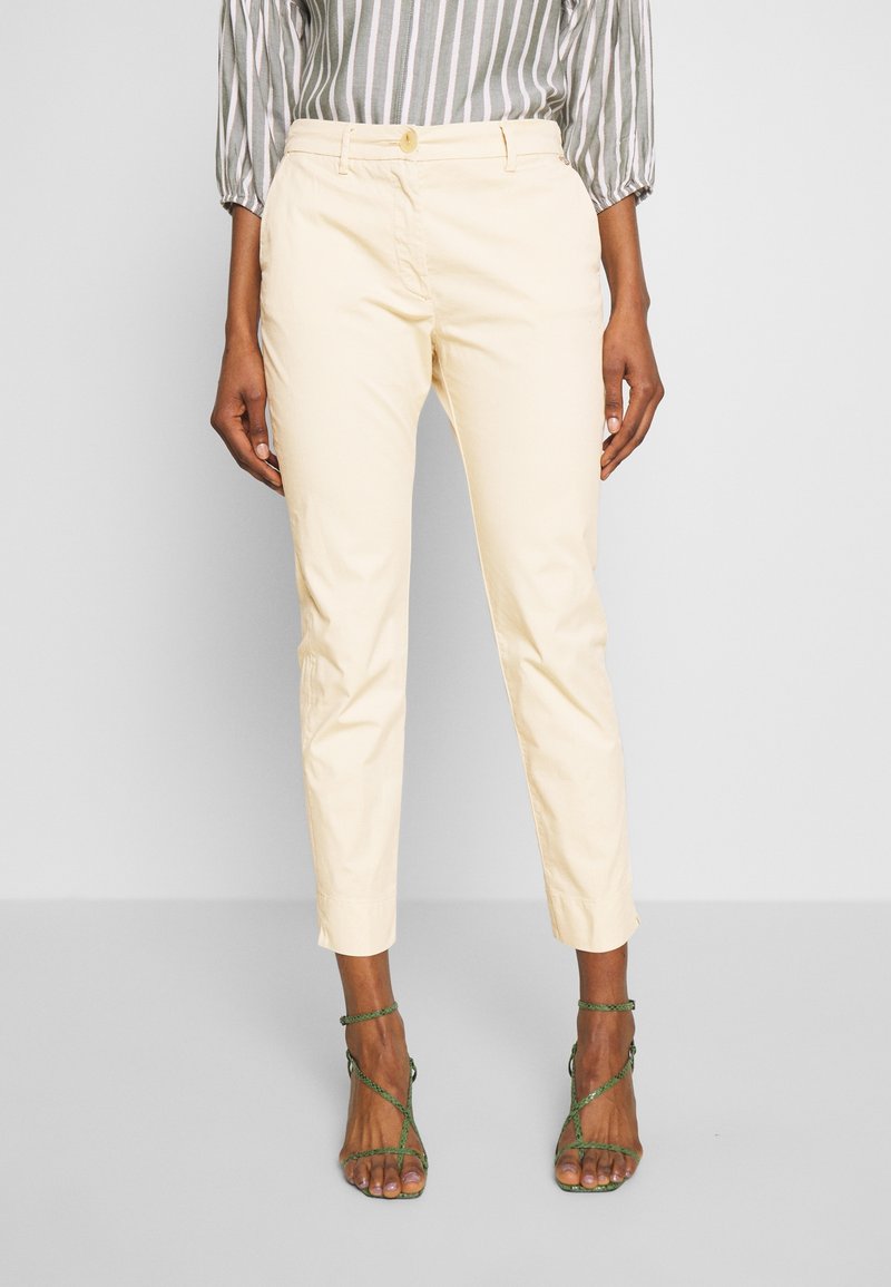 Kvinna klädd i beige beskurna byxor, gröna sandaler med klack och remmar, samt en grå och vit randig blus med trekvartsärmar.