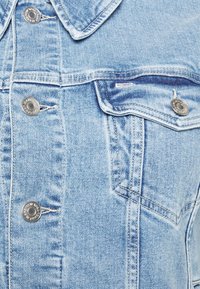 Chaqueta vaquera azul claro con cuello clásico, botones a presión plateados y dos bolsillos en el pecho. Presenta costuras visibles y un acabado suave y texturizado.