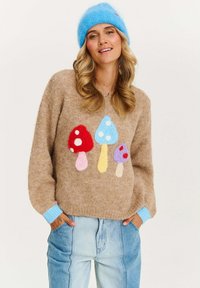 Sweter dzianinowy w kolorze jasnobrązowym z kolorowymi motywami grzybów. Posiada niebieskie ściągacze na rękawach i u dołu, luźny krój oraz miękką fakturę. Para dżinsów.