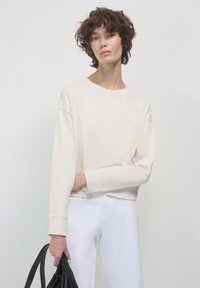 Felpa beige chiaro con collo rotondo e maniche lunghe, abbinata a pantaloni bianchi. La modella tiene una borsa nera, caratterizzata da una texture liscia.