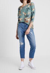 Bluse mit Blumenmuster in Teal, langärmlig; hellblaue, verwaschene Jeans; weiße Canvas-Sneaker. Lässiger Schnitt und schmal zulaufende Beinform.