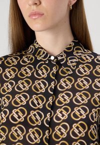 Blusa nera con un motivo geometrico ripetuto in oro, bianco e marrone. Presenta un colletto, chiusura con bottoni e una texture liscia.