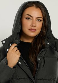 Chaqueta de plumas negra con capucha, con herrajes plateados, cierre de cremallera y textura acolchada. Lleva puesta sobre un suéter negro de punto.