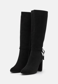 Par de botas de ante negras hasta la rodilla con tacones de bloque y correas decorativas que se envuelven, mostradas sobre un fondo blanco.