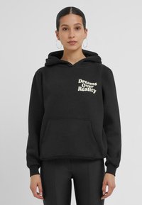 Zwarte hoodie met een voorzak. Kenmerkt een grafische tekstontwerp "Dreams Over Reality" in lichte kleur op de bovenste linkerkant. Zacht, textuurstof.