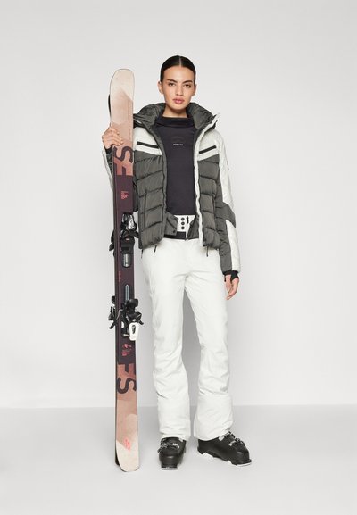 Ski-uitrusting: Puffjack in grijs en wit, zwarte basislaag, witte skibroek, zwarte skibotten, houdt beige skies vast met bindingaccenten.