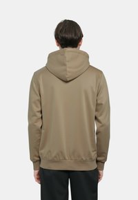 Giacca con cappuccio in tessuto color camoscio, caratterizzata da una texture liscia, polsini elastici e un orlo a coste. La vista posteriore mostra una silhouette aderente.