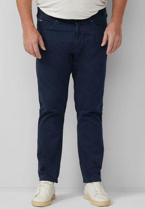 Man draagt donkerblauwe, slim-fit broek en witte sneakers, staand tegen een effen lichtgrijze achtergrond.