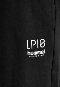 Sort træningstøj med struktureret stof. Med hvidt trykt logo "LP10" og "hummel SPORTSWEAR" langs det nedre område.