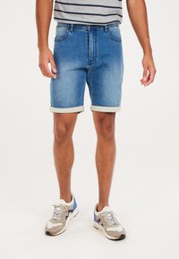 Pantalones cortos de mezclilla en azul claro con un acabado ligeramente desgastado, dobladillo vuelto y diseño de cinco bolsillos, combinados con zapatillas grises y una camisa a rayas.