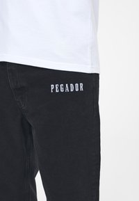 Svarta jeans med rak passform och synlig sömnad. "PEGADOR" är broderat i vitt på höger lår. Texturen verkar vara hållbar.