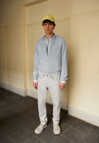 Lichtblauw overhem shirt over een grijs T-shirt, gecombineerd met lichtgrijze jeans en beige gewatteerde instap schoenen, afgerond met een gele pet.