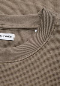 Κοντινό πλάνο γιακά πλεκτού πουκαμίσου σε χρώμα ταουπ με ριμπ ύφασμα στη λαιμόκοψη και μερικώς ορατή λευκή ετικέτα μάρκας.