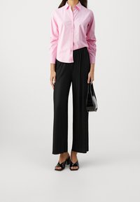 Henkilö, jolla on päällään vaaleanpunainen nappipaita, mustat pleatatut leveät housut, mustat korkeat sandaalit ja jota hän pitää mustaa käsilaukkua.