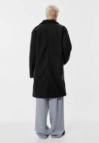 Zwarte jas met een gestructureerd ontwerp, voorzien van een kraag en een rechte snit. Gecombineerd met grijze, loszittende joggingbroeken.