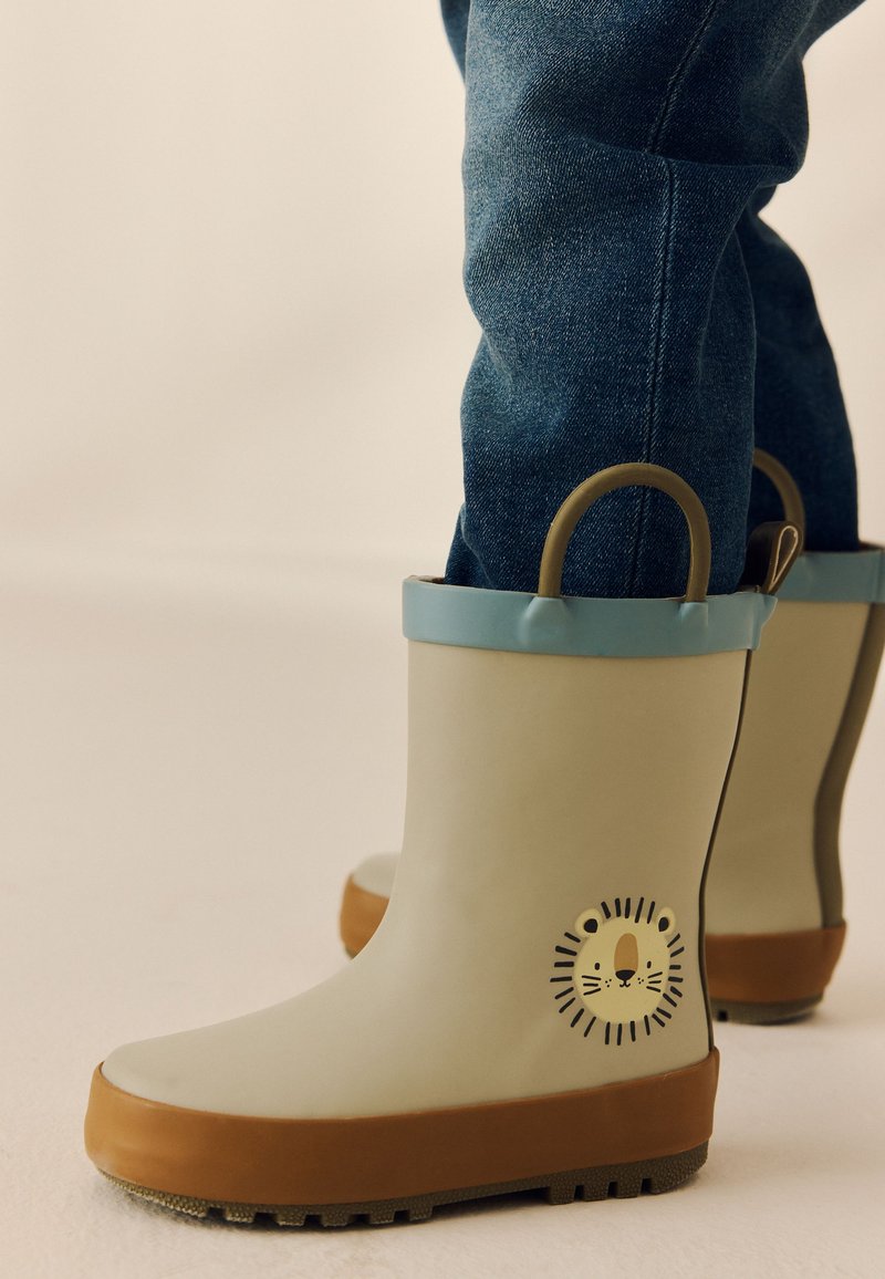 Bambino che indossa stivali da pioggia beige con bordo blu e disegno di faccia di leone, abbinati a jeans blu.
