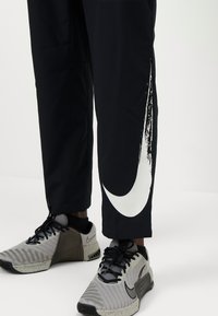 Sorte træningsbukser har et stort hvidt Nike swoosh med et tekstureret design på siden. Bæres med grå og sorte mønstrede sneakers.