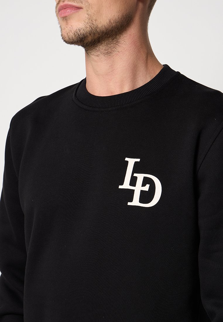 Mann in schwarzer Sweatshirt mit weißem "LD"-Logo auf der linken Brust, sichtbar von Nacken bis zur Mitte des Oberkörpers vor einem einfarbigen Hintergrund.