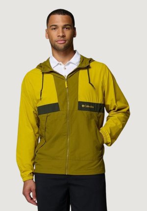Homme portant une veste zippée Columbia jaune et vert olive avec capuche, une chemise blanche à col, et un pantalon noir, debout avec une main dans la poche.
