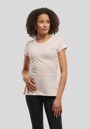 Femme aux cheveux bouclés portant un t-shirt rose clair avec un petit motif de visage de chien et des leggings noirs, debout devant un fond gris.