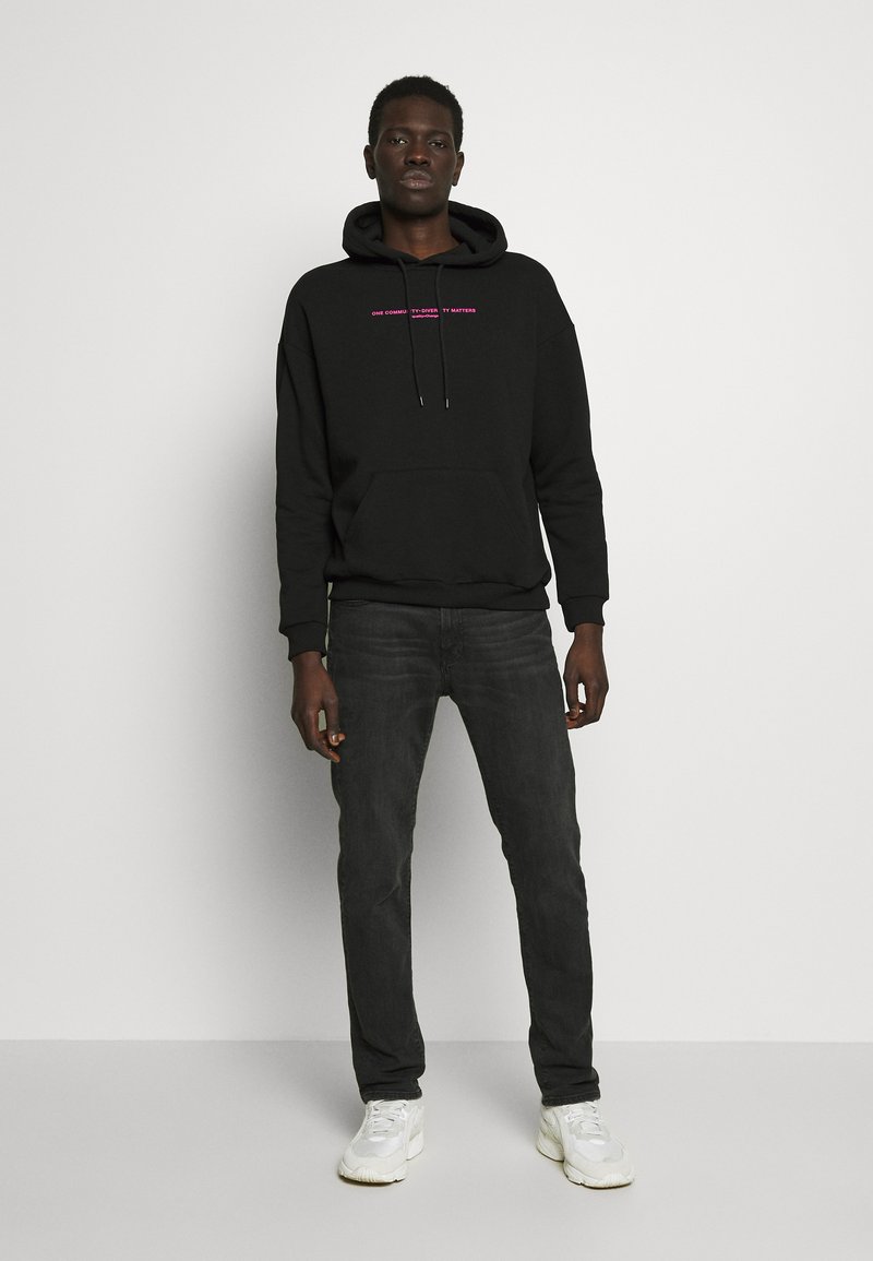 Black hoodie with pink text, featuring a kangaroo pocket and drawstring. Paired with dark gray jeans and white sneakers.