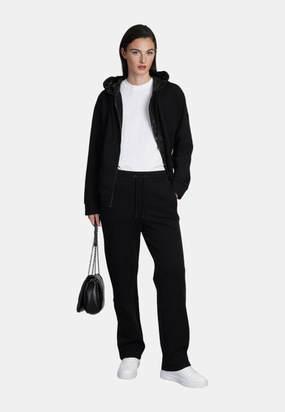 Zwarte zip-up hoodie met een trekkoordcapuchon, gecombineerd met zwarte joggingsbroek. Een witte T-shirt eronder, witte sneakers en een zwarte handtas.