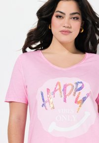 Camiseta de manga corta rosa con escote en V, que presenta un gráfico de lentejuelas con la palabra "HAPPY" y el texto "VIBES ONLY" en blanco, sobre una suave tela de algodón.