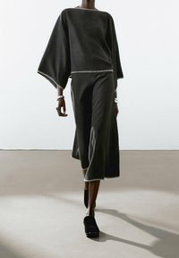 Tenue noire avec un haut large et carré et une jupe évasée jusqu'à la cheville, toutes deux ornées de surpiqûres blanches contrastantes. Le mannequin porte des chaussures noires à enfiler.