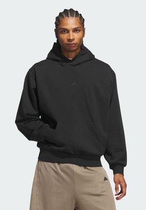 Mand med flettet hår iført en sort hættetrøje og beige strukturerede shorts, stående med den ene hånd i hættetrøjens lomme mod en ensfarvet baggrund.