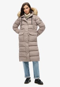 Manteau long et bouffant de couleur beige avec une capuche doublée de fourrure, poches avant et fermeture éclair. Porté par-dessus un col roulé crème et un jean bleu. Chaussures noires.