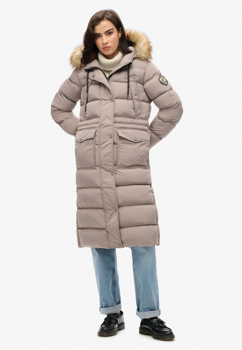 Manteau long et bouffant de couleur beige avec une capuche doublée de fourrure, poches avant et fermeture éclair. Porté par-dessus un col roulé crème et un jean bleu. Chaussures noires.