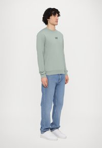 Felpa a maniche lunghe verde chiaro con scollo rotondo e logo sul petto, abbinata a jeans azzurri e sneakers bianche.